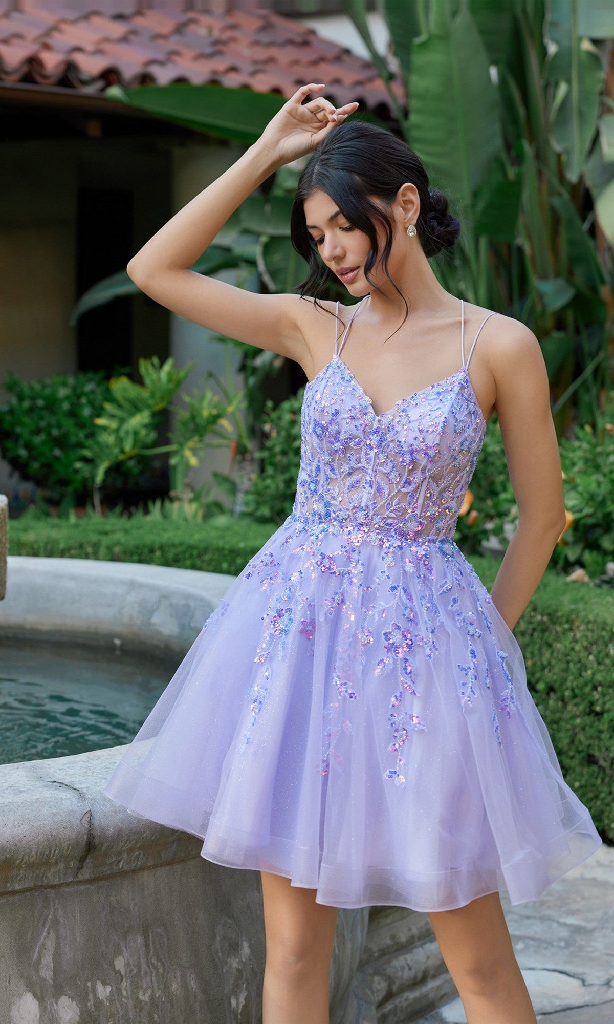 A woman stands outdoors by a fountain, surrounded by lush greenery, wearing the Short Homecoming Dress: Juliet JT988N in lilac. The dress features a sequin-embellished corset bodice that catches the light and is complemented by an elegant fit-and-flare tulle skirt.